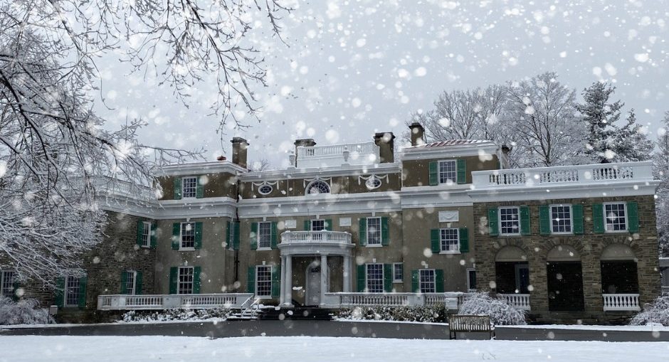 A large, historic mansion with green shutters and snow-covered grounds is seen under falling snowflakes, surrounded by leafless trees on a snowy winter day.