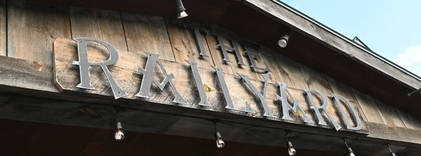 Large metal letters spelling THE RAILYARD are mounted on weathered wooden boards above a building entrance, with exposed light bulbs hanging below—reminiscent of Burleigh’s Luncheonette—set against a blue sky.