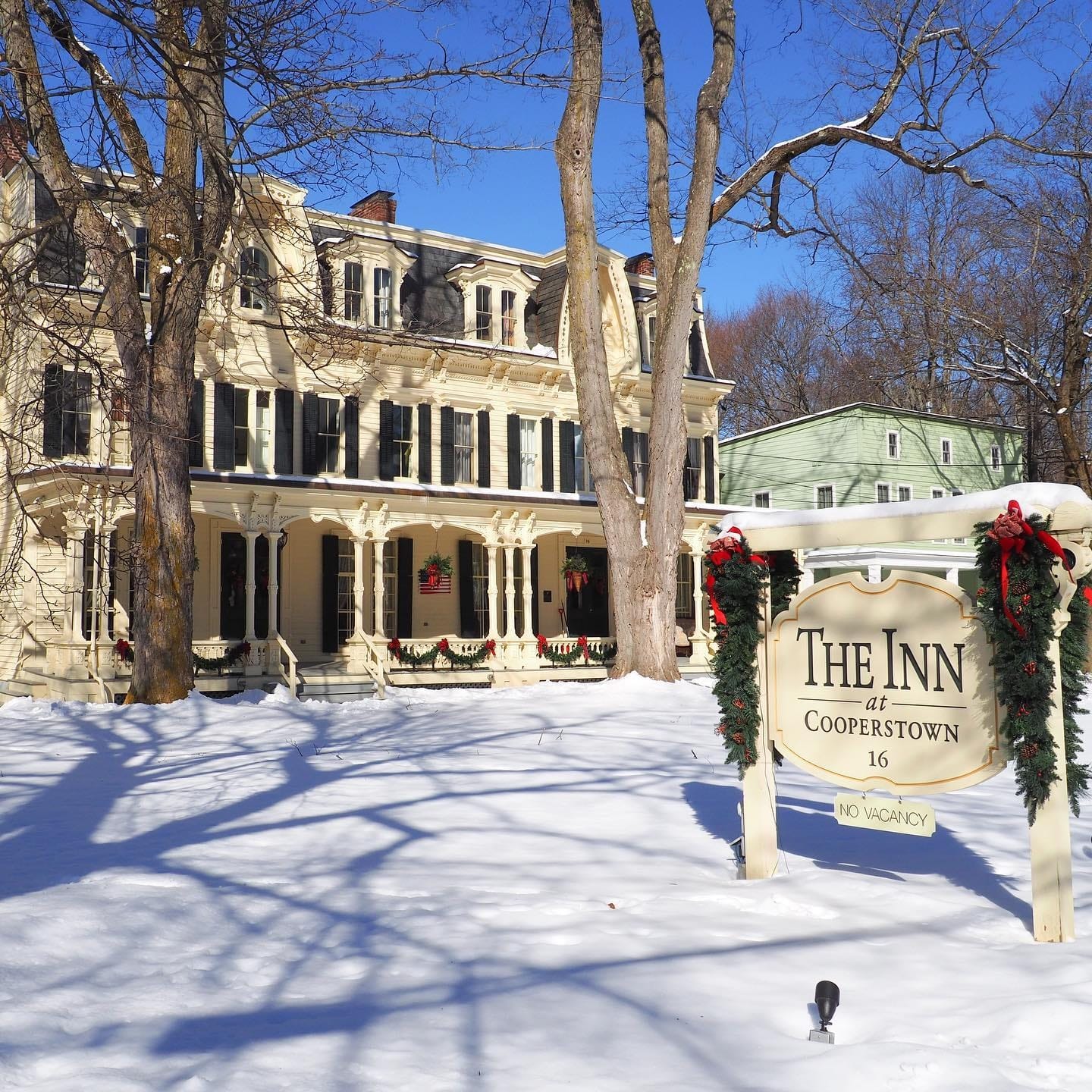 The historic Inn at Cooperstown, adorned with festive holiday decorations, stands behind a snow-covered yard and its welcoming sign on a bright, sunny winter day.