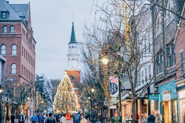 A festive city street lined with shops and trees adorned with holiday lights, people strolling past Lang House, and a large decorated Christmas tree in front of a church with a tall steeple in the background.