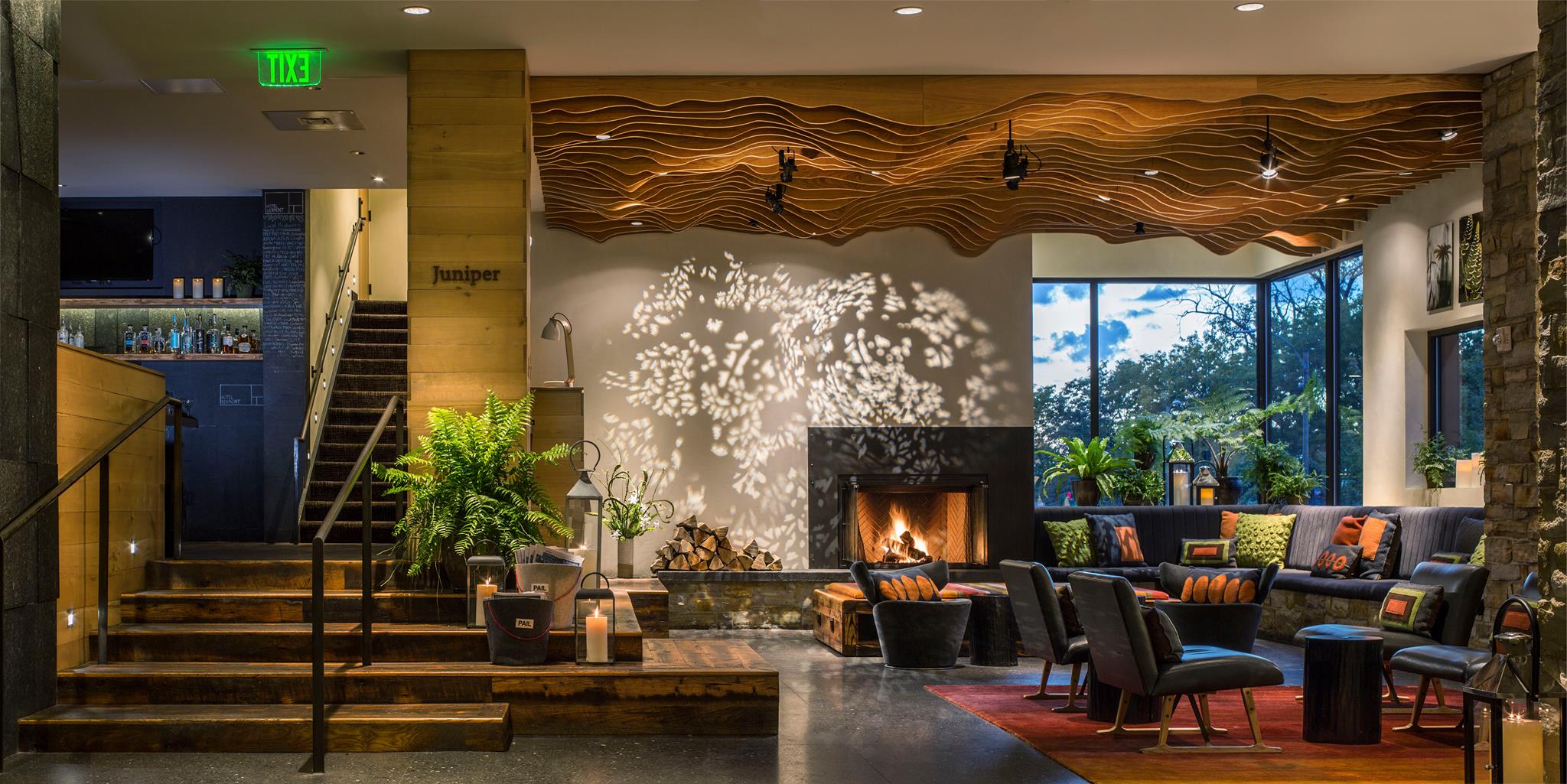 A modern Hotel Vermont lobby with warm lighting, a lit fireplace, black sofas with colorful pillows, indoor plants, and a wavy wooden ceiling feature. Shadows of tree branches are cast on the wall, creating a cozy atmosphere.