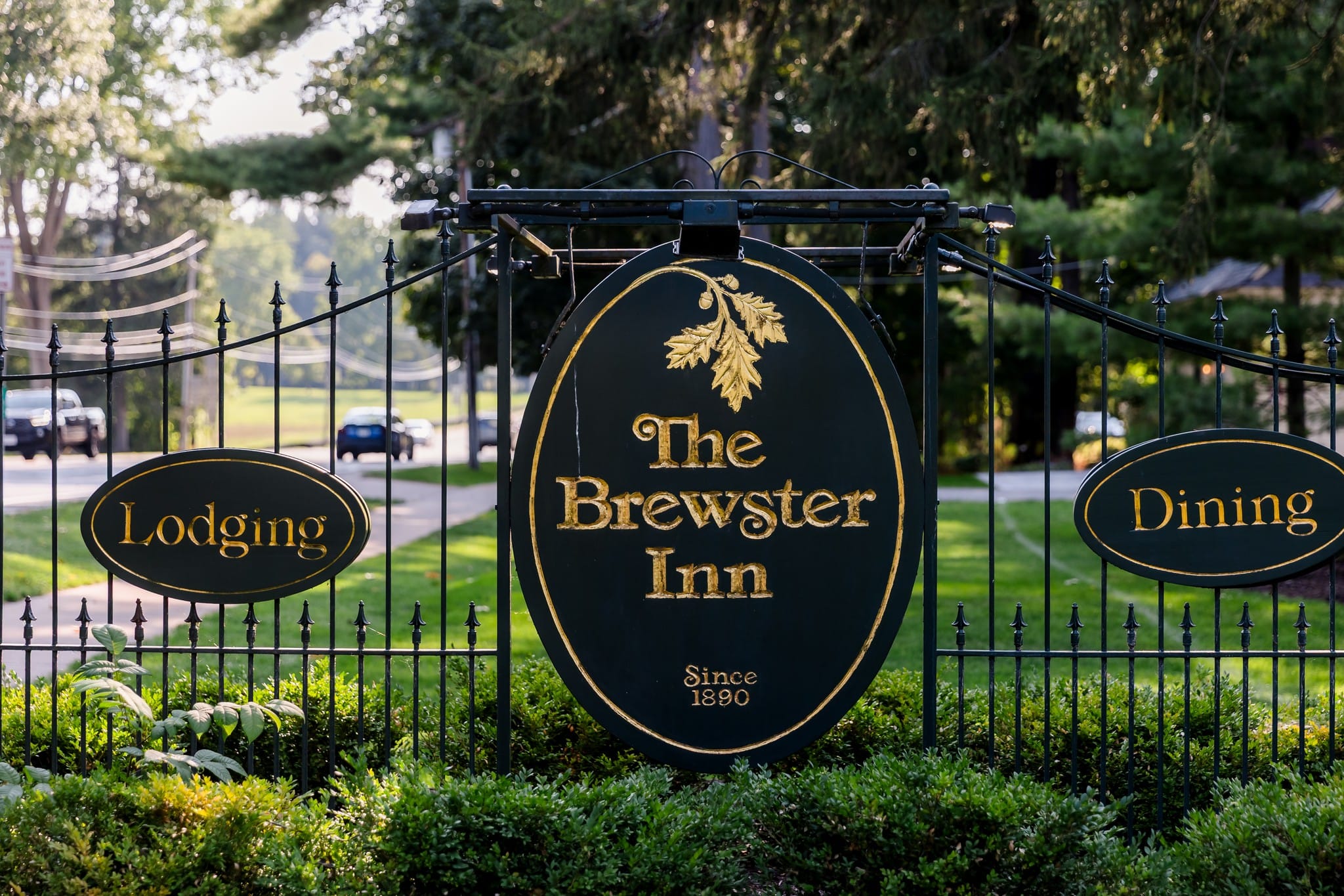 A black and gold sign for The Brewster Inn, Since 1890, features smaller signs for Lodging and Dining, all attached to a black metal fence with trees and a lawn enhancing the historic charm in the background.
