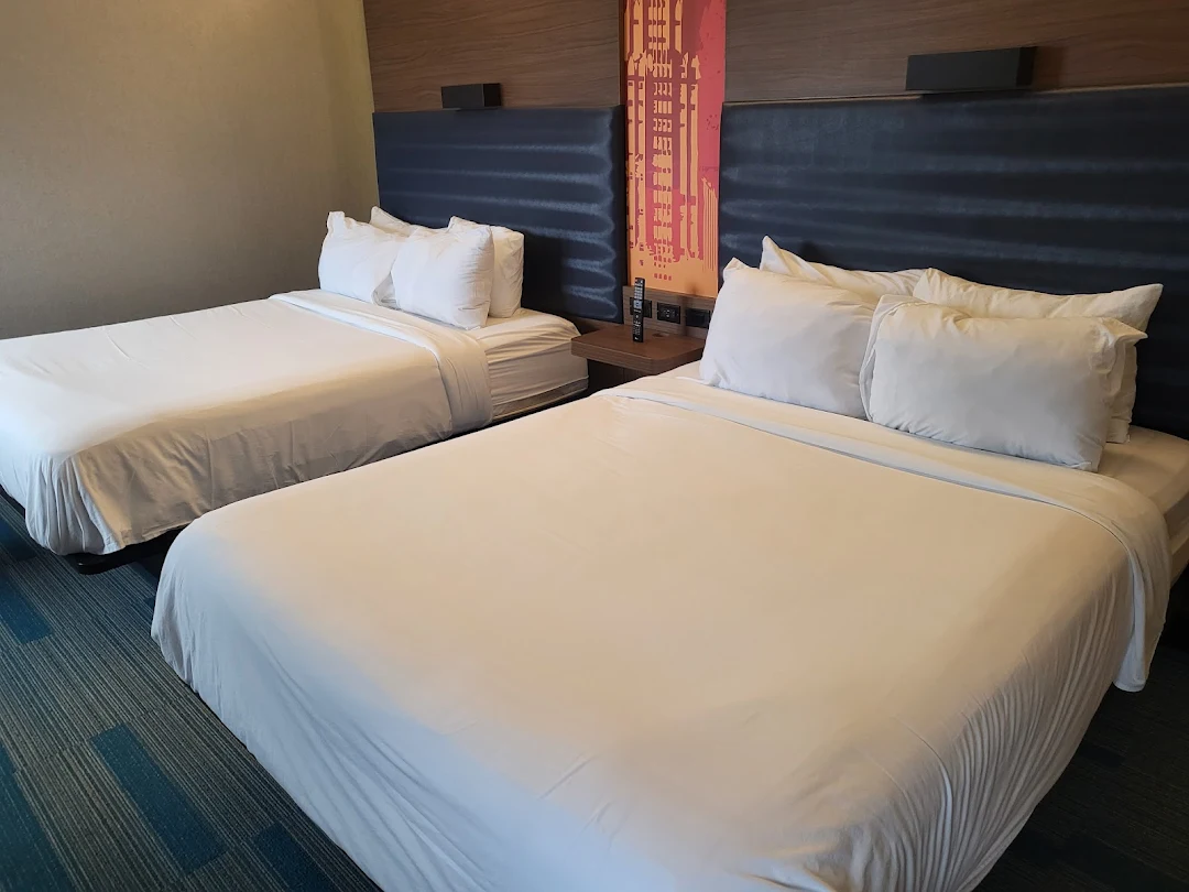 Two neatly made beds with white linens and pillows in a modern Aloft Syracuse hotel room, featuring a blue padded headboard, wooden accents, bedside tables, and a colorful wall decoration.
