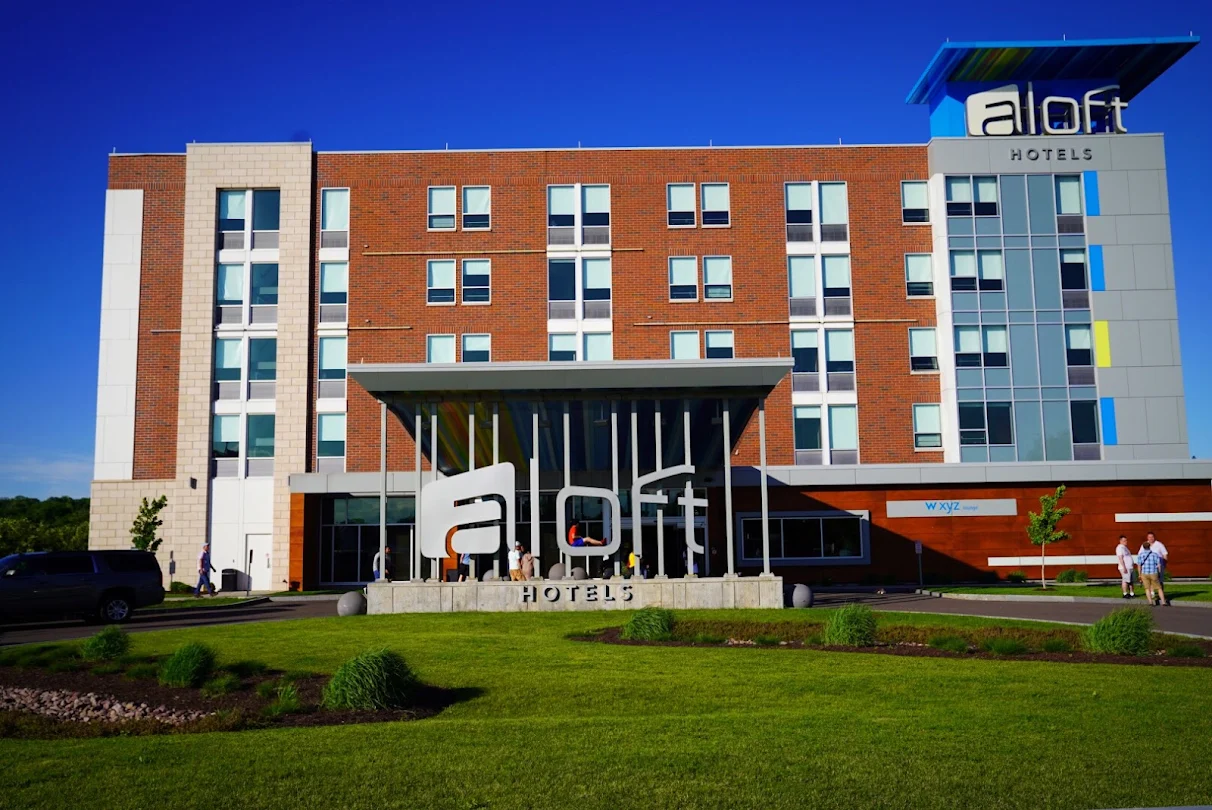 A modern Aloft Syracuse building with large windows and a bold sign out front, surrounded by green grass and landscaping beneath a clear blue sky. A few people are walking near the entrance.