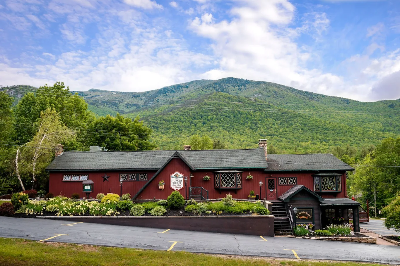 A rustic red building with a dark roof sits beside a sloped parking lot at Hungry Trout Resort, surrounded by lush green trees and flowering shrubs, with forested mountains and a partly cloudy sky in the background.