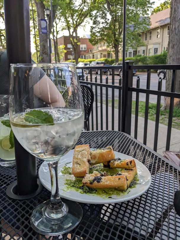 A glass of sparkling water with mint and a plate of toasted bread with pesto sit on a black metal patio table outside The Classic Kitchen and Cocktails, with trees, houses, and a street visible in the background.