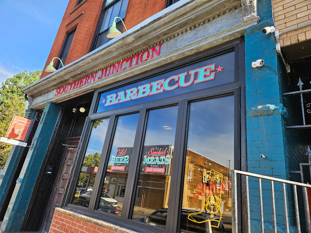 The storefront of Southern Junction Barbecue features large windows reflecting the street, a neon sign, and a colorful BARBECUE sign above the entrance on this classic brick building.