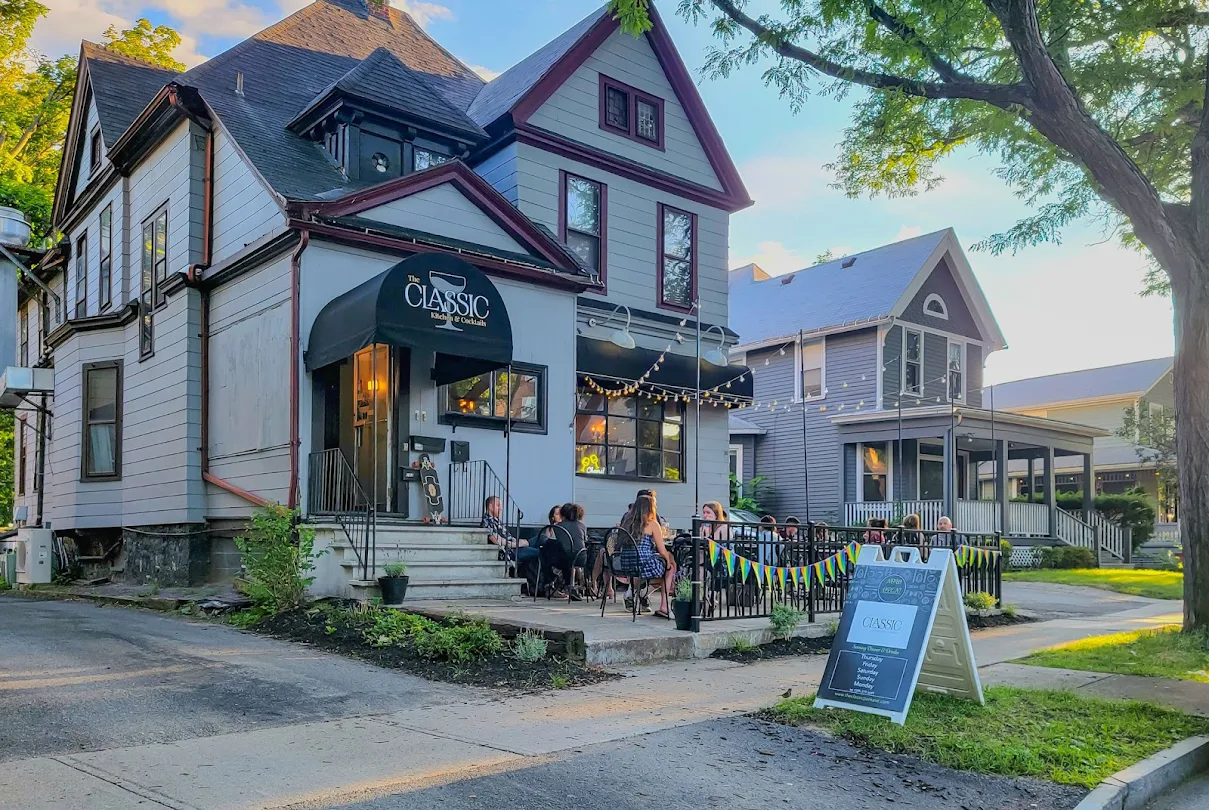 People sit at outdoor tables in front of The Classic Kitchen and Cocktails, a cozy café in a renovated house on a tree-lined street. A sign near the sidewalk reads Classic, with nearby houses and greenery visible under a bright sky.