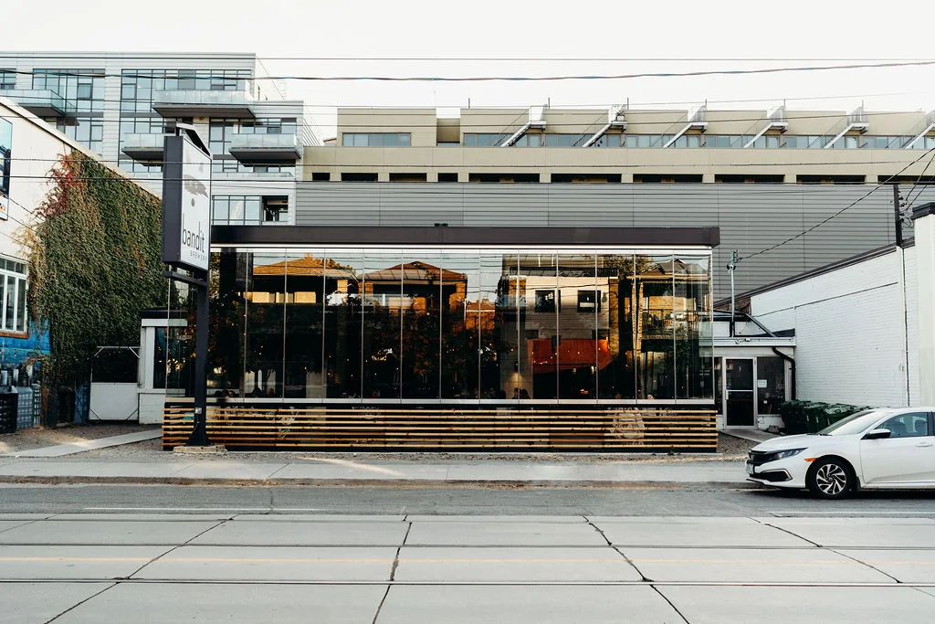 A modern building with large glass windows, reflecting nearby structures, sits along a street with a white car parked in front. Home to Bandit Brewery, it features horizontal wooden slats and blends urban vibes with touches of greenery.