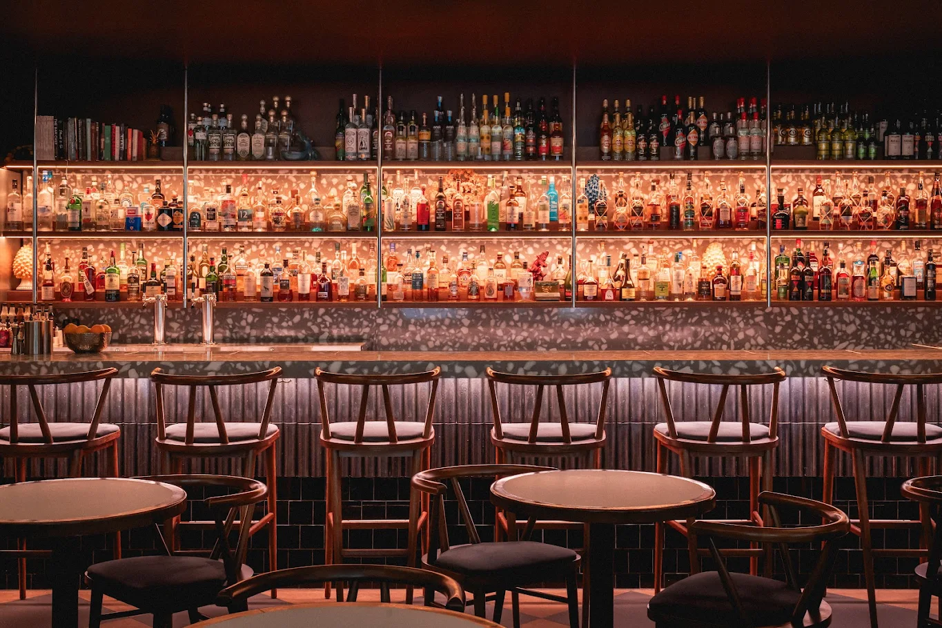 A stylish, dimly lit bar—Bar Le Mal Nécessaire—features rows of empty wooden chairs facing a long counter. Behind the counter, shelves of liquor glow with warm lighting, while round tables invite guests in the foreground.