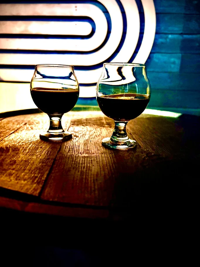 Two glasses of dark liquid sit on a round wooden table, with a blue wall and white curved line pattern in the background. The moody, artistic atmosphere hints at the craftsmanship found at Brasserie Distillerie Beauregard.