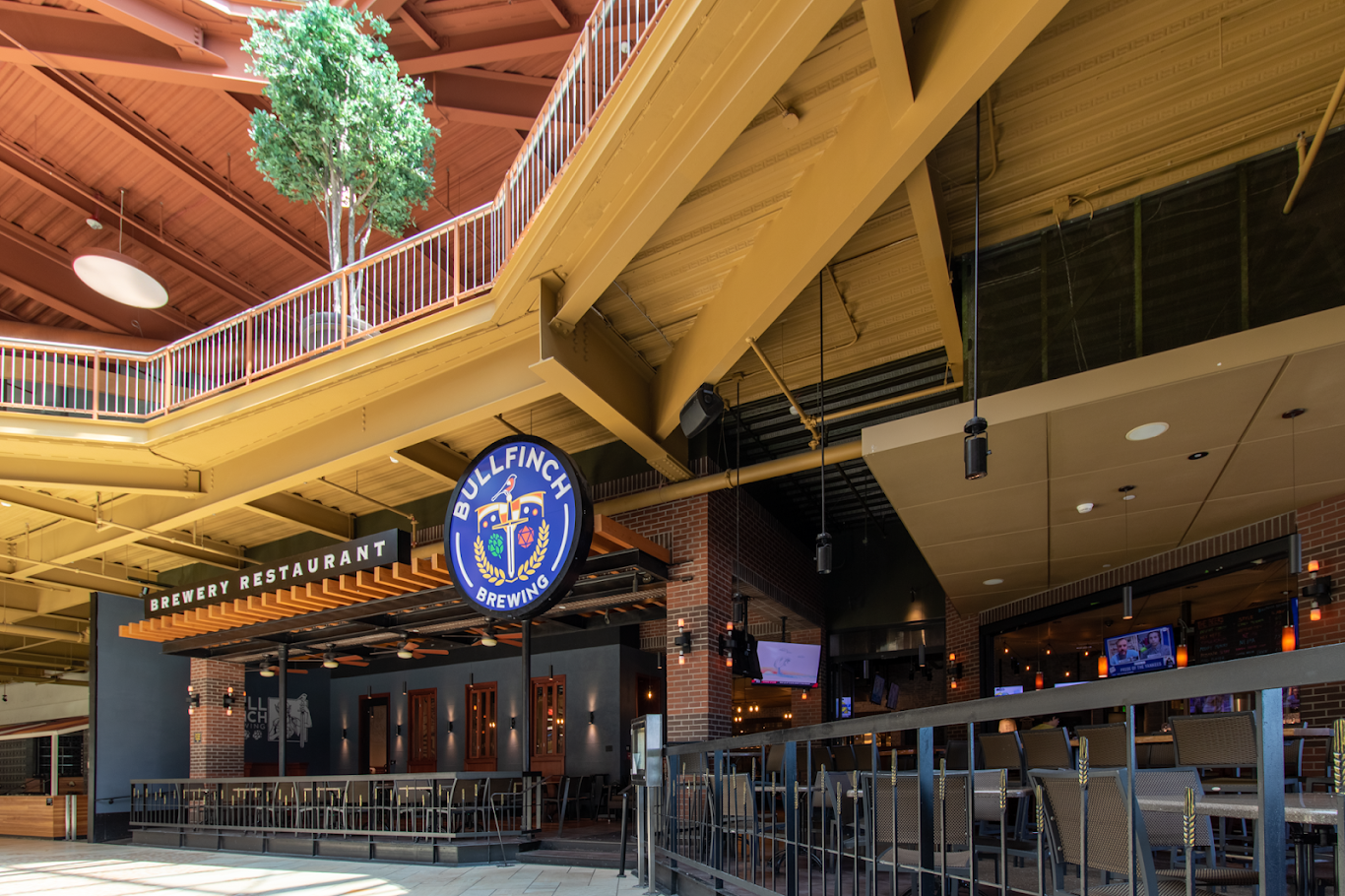 A spacious indoor brewery restaurant, Bullfinch Brewpub features yellow metal beams, an upper walkway, a Bullfinch Brewery sign, and a bar area with chairs and TV screens. A potted tree on the upper level adds a refreshing touch of greenery.