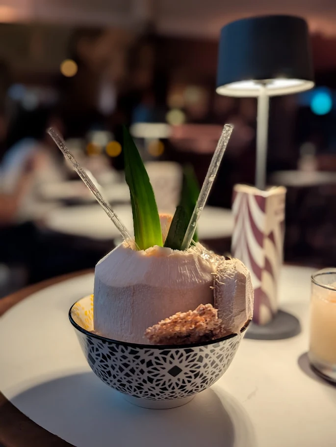 A coconut with two straws, garnished with green leaves and dried fruit, is served in a decorative bowl on a round table at Bar Le Mal Nécessaire, with a lamp and a drink in the background.