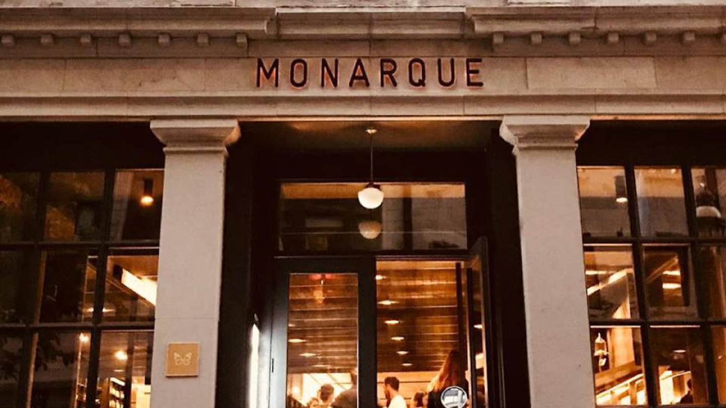 Street view of Monarque restaurant's entrance, featuring large glass windows, double doors, and the Monarque name in bold letters above. Warm lighting glows from inside, where people can be seen enjoying their evening.