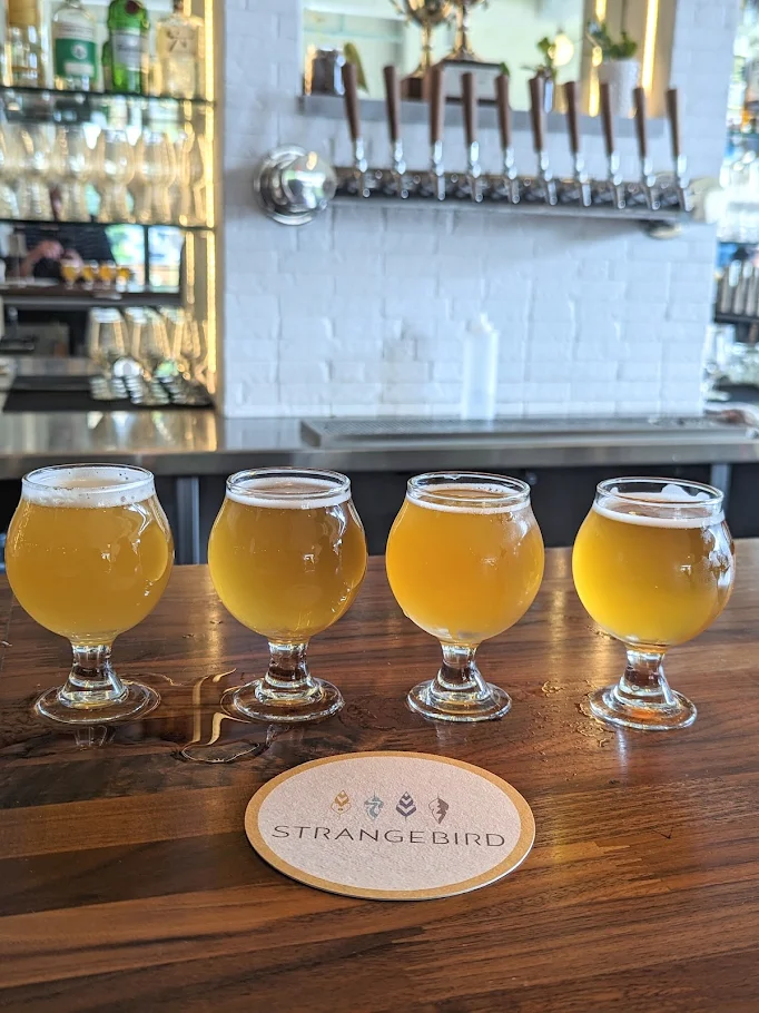 Four small glasses of beer from Strangebird are lined up on a wooden bar in front of a branded coaster. Behind them, beer taps and shelves with bottles decorate the white brick wall.