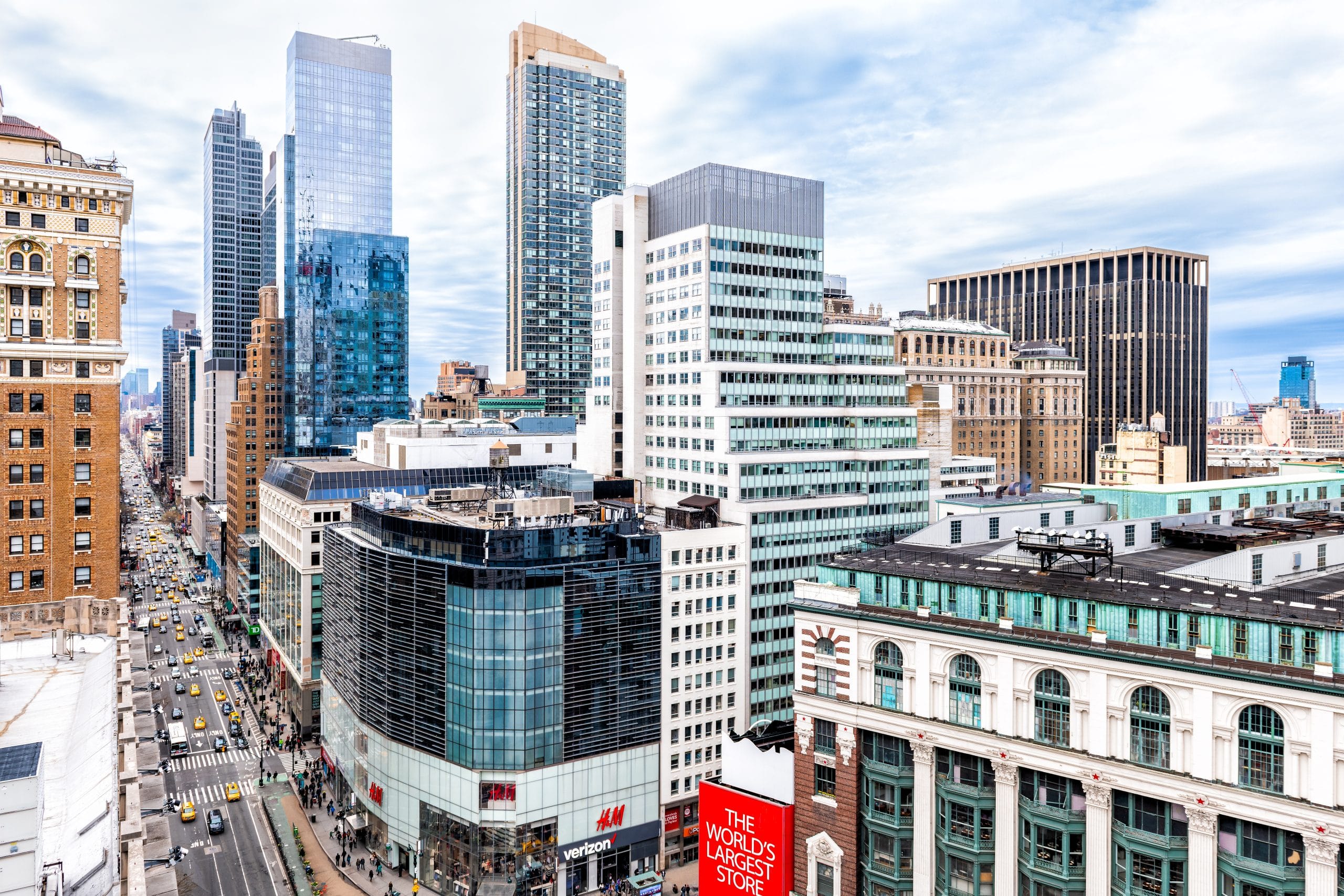 A busy New York City street near Herald Square, close to Amtrak Penn Station, lined with tall buildings—modern glass skyscrapers and older brick structures—with yellow taxis below and a red sign for The World's Largest Store visible.