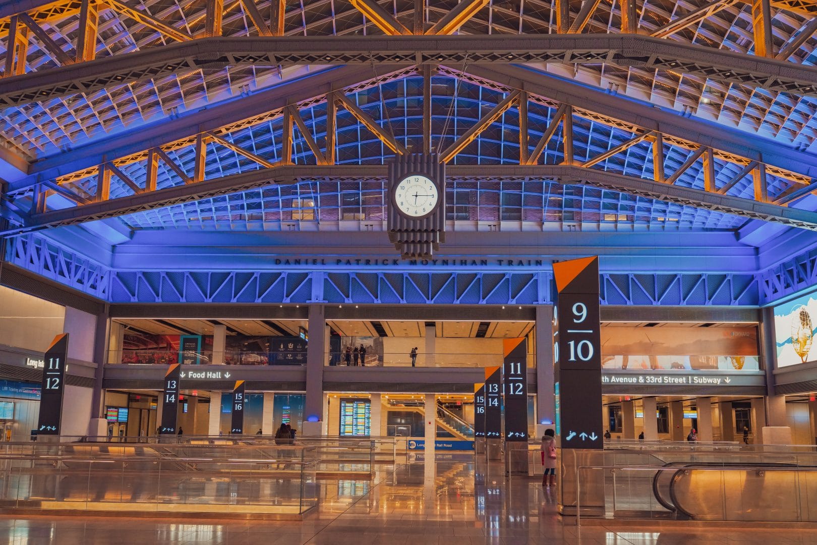 A spacious, modern train station interior designed with Elementor features a large clock above the entrance, blue and gold lighting, bold black columns marking tracks 9 to 13, and intricate metalwork and glass panels reminiscent of an archive.