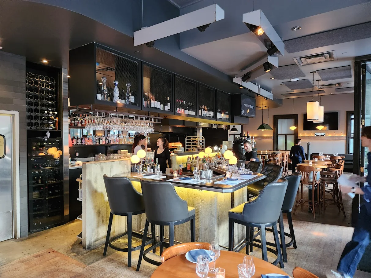 A modern restaurant bar with high chairs around a counter, glassware and bottles on shelves, and several people sitting or working behind the bar. The stylish, warmly lit atmosphere gives Pubjelly its inviting charm.