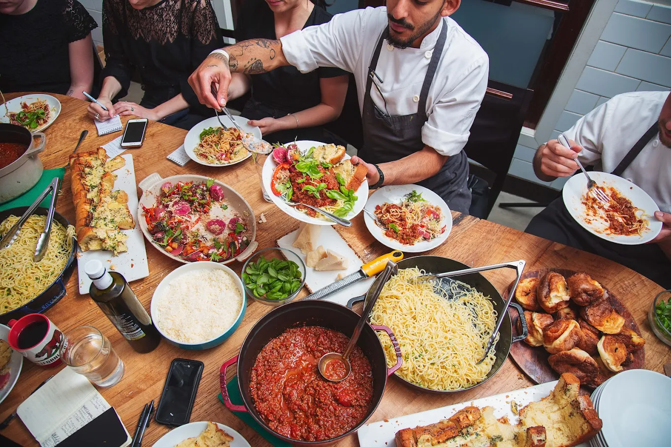 A group of people sit around a wooden table at Richmond Station, enjoying pasta dishes, sauce, salad, bread, and drinks as a chef serves food onto plates.