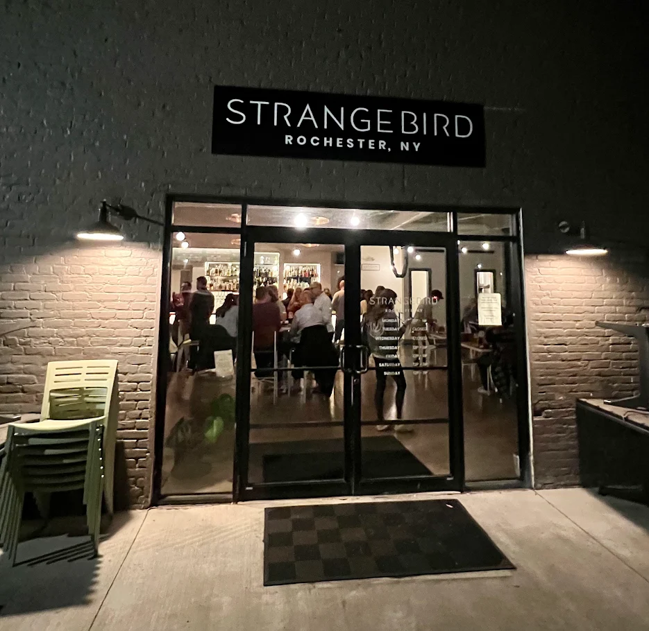 Exterior view of Strangebird, a popular spot in Rochester, NY. The entrance features glass double doors, a black Strangebird sign above, stacked chairs to the left, and people visible inside enjoying warm lighting.