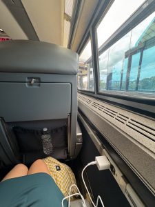 A mother and daughter sit by an Amtrak train window with their legs visible; a patterned bag rests below, a water bottle is tucked in the seat pocket, and a phone charges from a nearby outlet during their train trip.