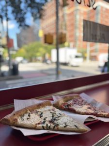 Two slices of pizza on paper plates rest on a red counter by a window, with a blurred city street and brick buildings visible outside in the background—a perfect pit stop for a Mother Daughter Amtrak Train Trip through the city.