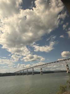 A long steel bridge spans across a wide river under a sky filled with large, fluffy clouds. Trees line the distant shore, sunlight brightens the scene, and an Amtrak train glides across the bridge—perhaps carrying families on a memorable mother-daughter train trip.