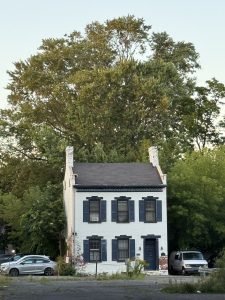 A small white brick house with blue shutters stands in front of leafy trees, evoking memories of a cozy stop on a Mother Daughter Amtrak Train Trip. Two chimneys lean outward, and two cars are parked on either side. The house has visible peeling paint near its foundation.