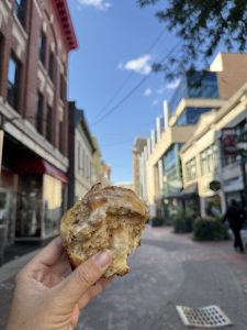A hand holds a partially eaten cinnamon roll in focus, capturing a sweet moment from a Mother Daughter Amtrak Train Trip, with a blurred urban street scene and brick buildings in the background under a blue sky.