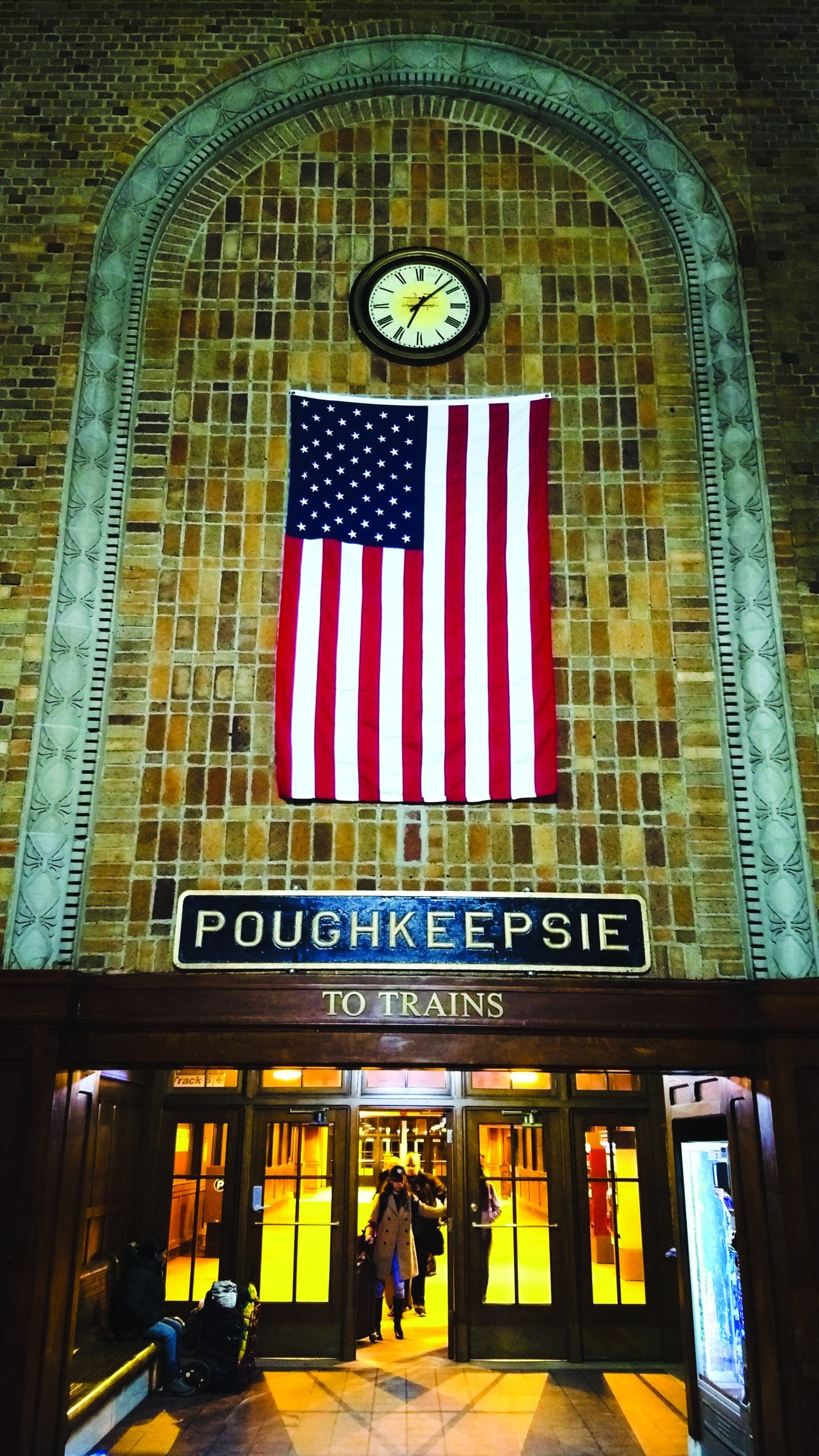 A large American flag and a clock hang above the “POUGHKEEPSIE TO TRAINS” sign in a brick-walled Amtrak station, where Majors In Success come and go—people are visible entering and exiting through doors below.