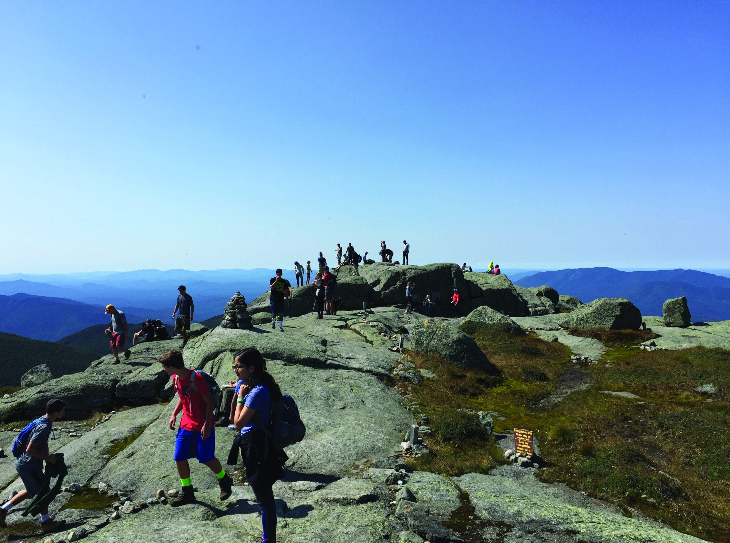 A group of hikers walk along a rocky mountain summit under a clear blue sky, embracing the spirit of True Great Adventures, with distant mountain ranges visible in the background.