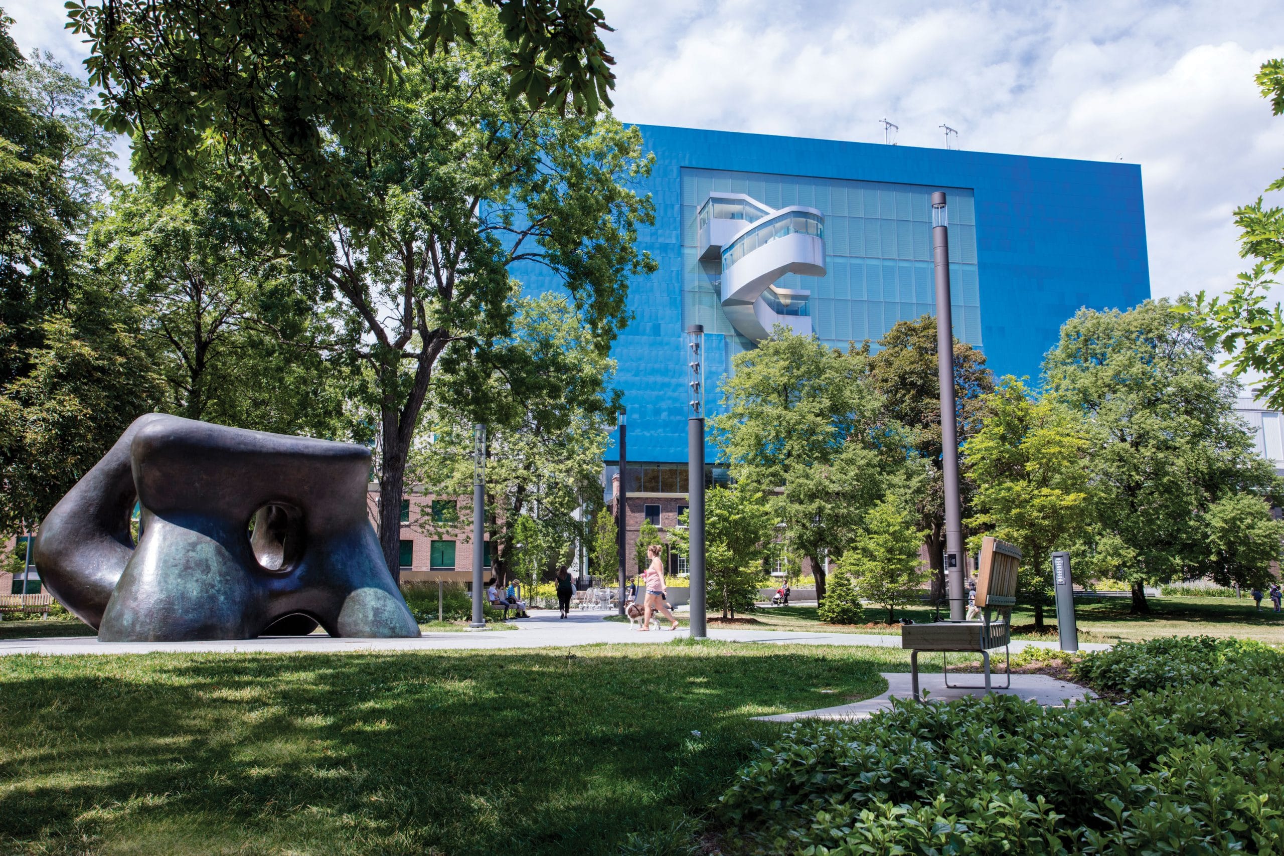 A modern blue glass building with a unique silver architectural feature stands amid trees and greenery. In the foreground, an abstract black sculpture and a few people walking reflect the creative energy of Toronto's Arts Scene.