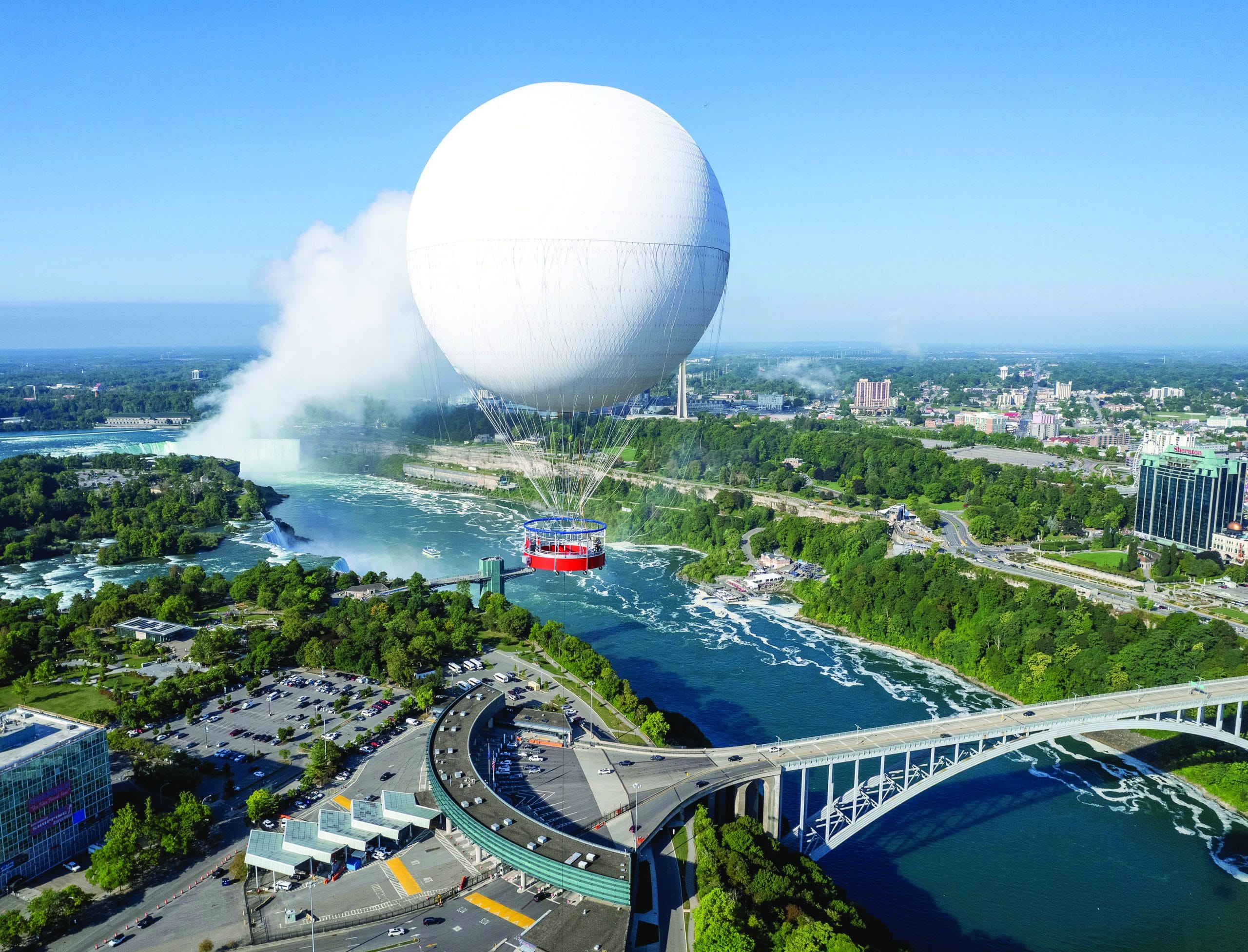 A large white tethered balloon carrying a red gondola floats above a river near a bridge, while people enjoy power walking along the riverside path with mist rising from Niagara Falls and a city surrounded by green trees in the background.