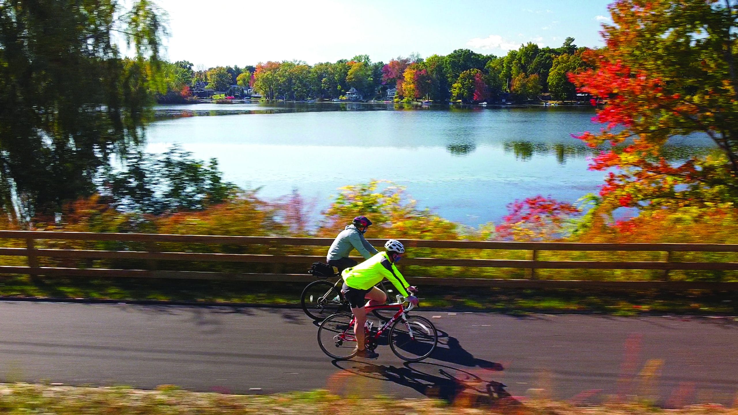 Two cyclists ride on a paved trail beside a lake, bordered by a wooden fence and colorful autumn trees—a peaceful Out of Doors scene perfect for those seeking True Great Adventures. The water reflects the blue sky and foliage beautifully.