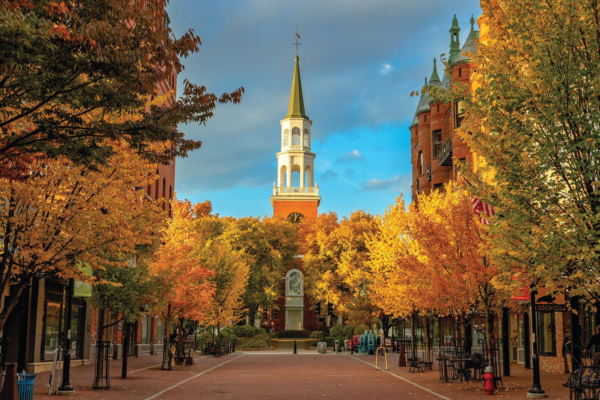 A pedestrian street in Burlington, Vermont, lined with autumn trees in vibrant orange and yellow, leads to a white church steeple under a partly cloudy sky. Historic brick buildings and outdoor seating frame this charming voyage.