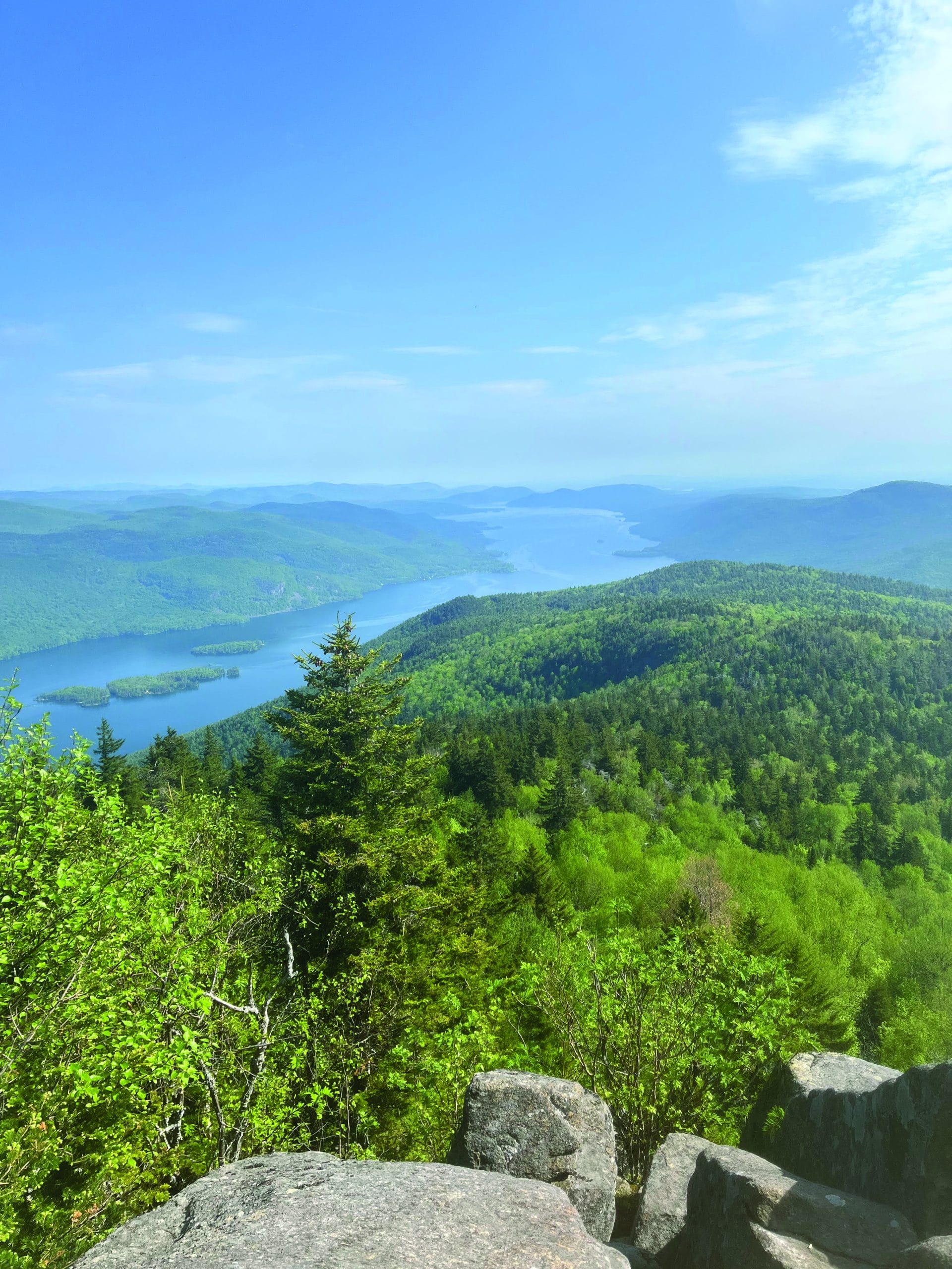 View from a mountaintop showing lush green forests, scattered trees, and a winding blue river stretching into the distance under a bright, partly cloudy sky. Large rocks rest in the foreground—perfect for those seeking True Great Adventures out of doors.