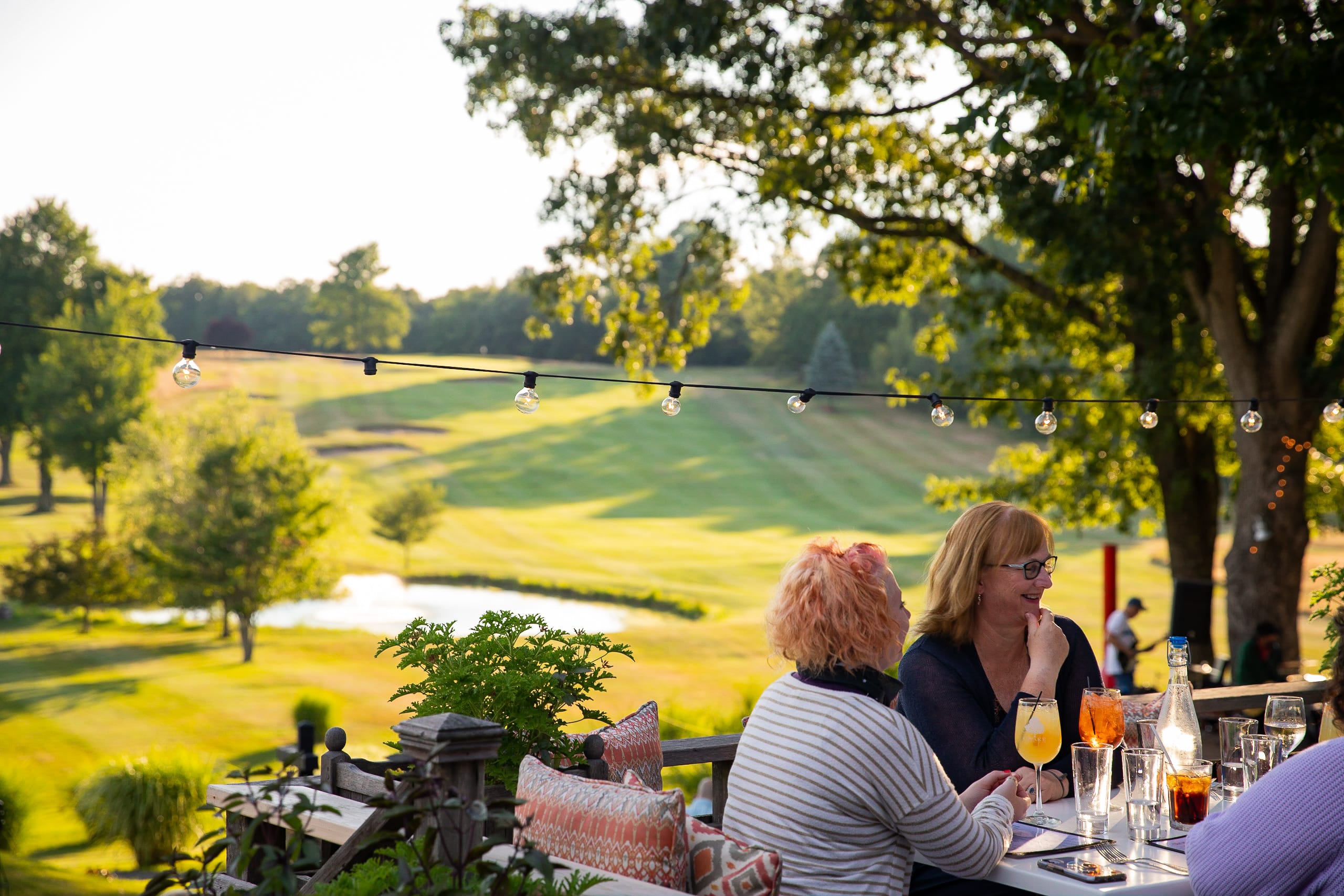 Two women sit at an outdoor patio table at Farmhouse Tap & Grill, enjoying drinks and conversation, overlooking a lush, sunlit golf course with trees and string lights hanging above.