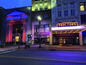 A brightly lit theater marquee reading PROCTORS glows in yellow and purple lights at dusk, with nearby buildings illuminated by colorful lights. A quiet, empty street in front awaits the bustle of power walking patrons arriving for the evening show.