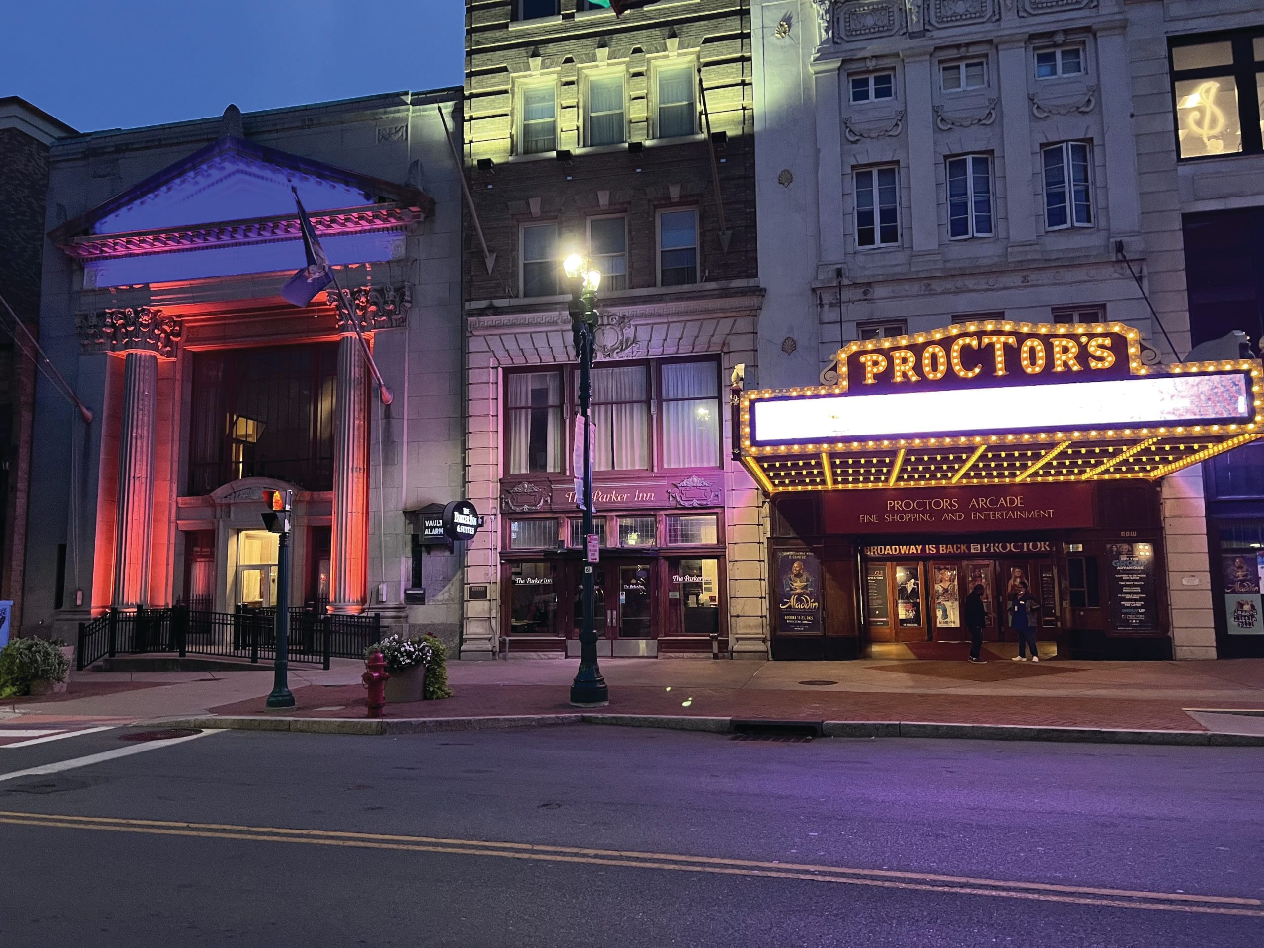 A brightly lit theater marquee reading PROCTORS glows in yellow and purple lights at dusk, with nearby buildings illuminated by colorful lights. A quiet, empty street in front awaits the bustle of power walking patrons arriving for the evening show.