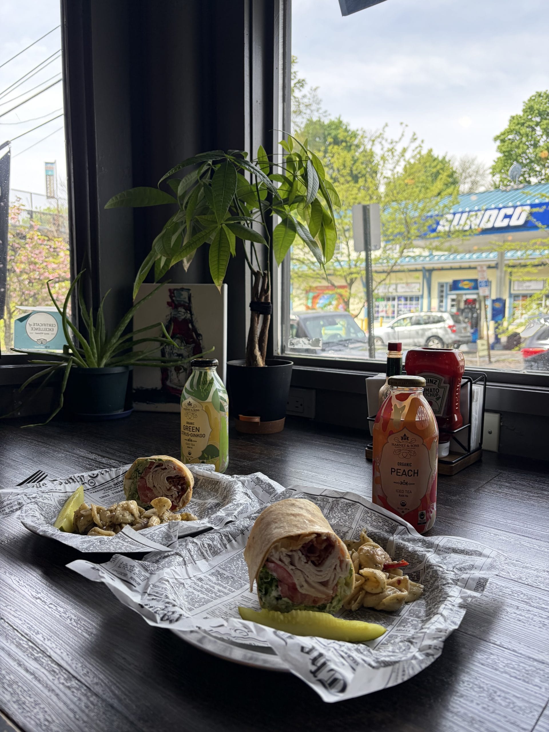 Two deli wraps with pickles and chips sit by a window, ready to enjoy after a Walking Tour of Poughkeepsie, alongside bottled drinks, condiments, a small plant, and a street view of cars and a Sunoco gas station outside.