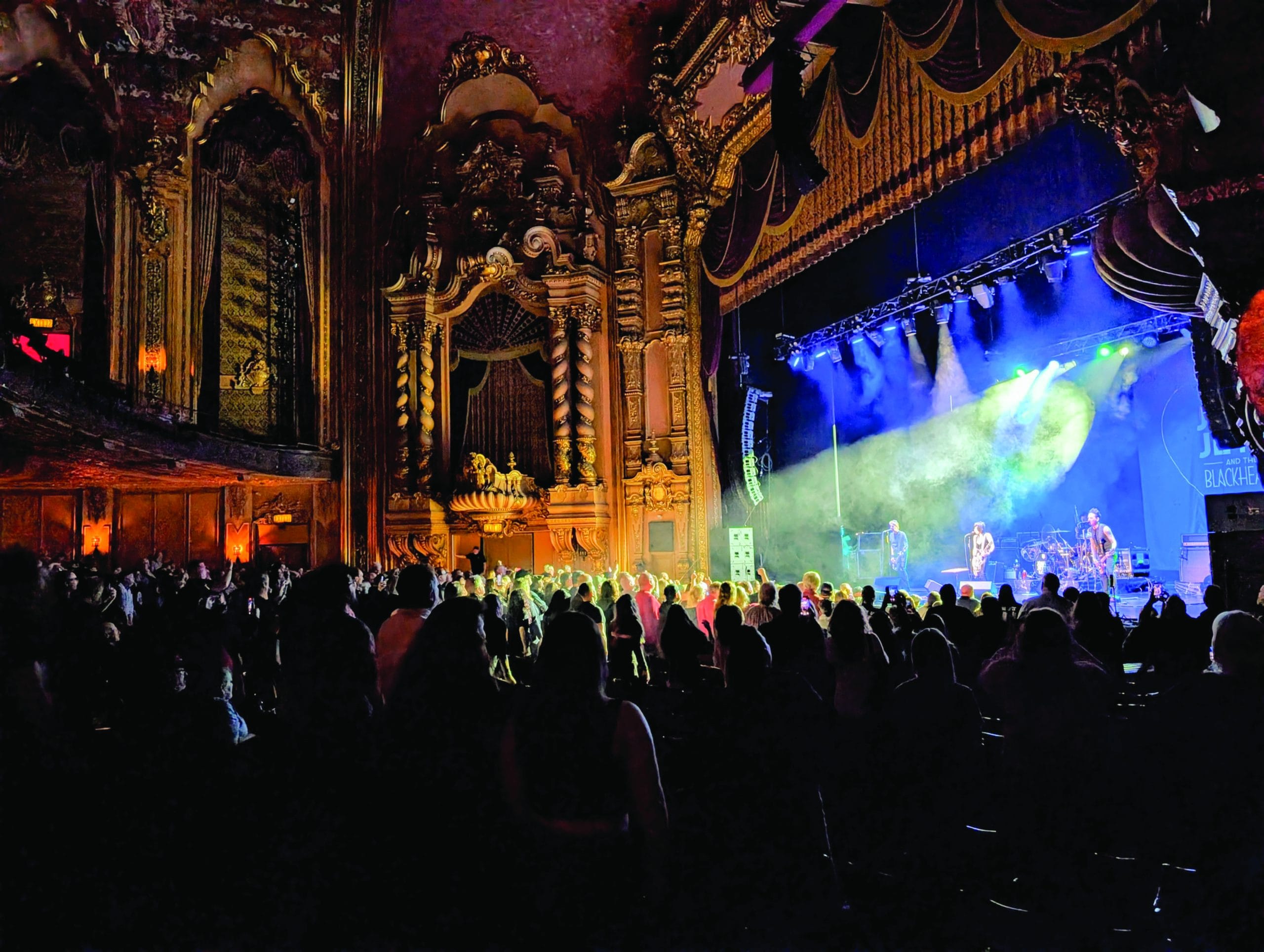 A crowd watches a live concert in a grand, ornately decorated theater with tall arches and gold details, some fans energized from power walking to the venue as dramatic stage lighting illuminates the performing band.