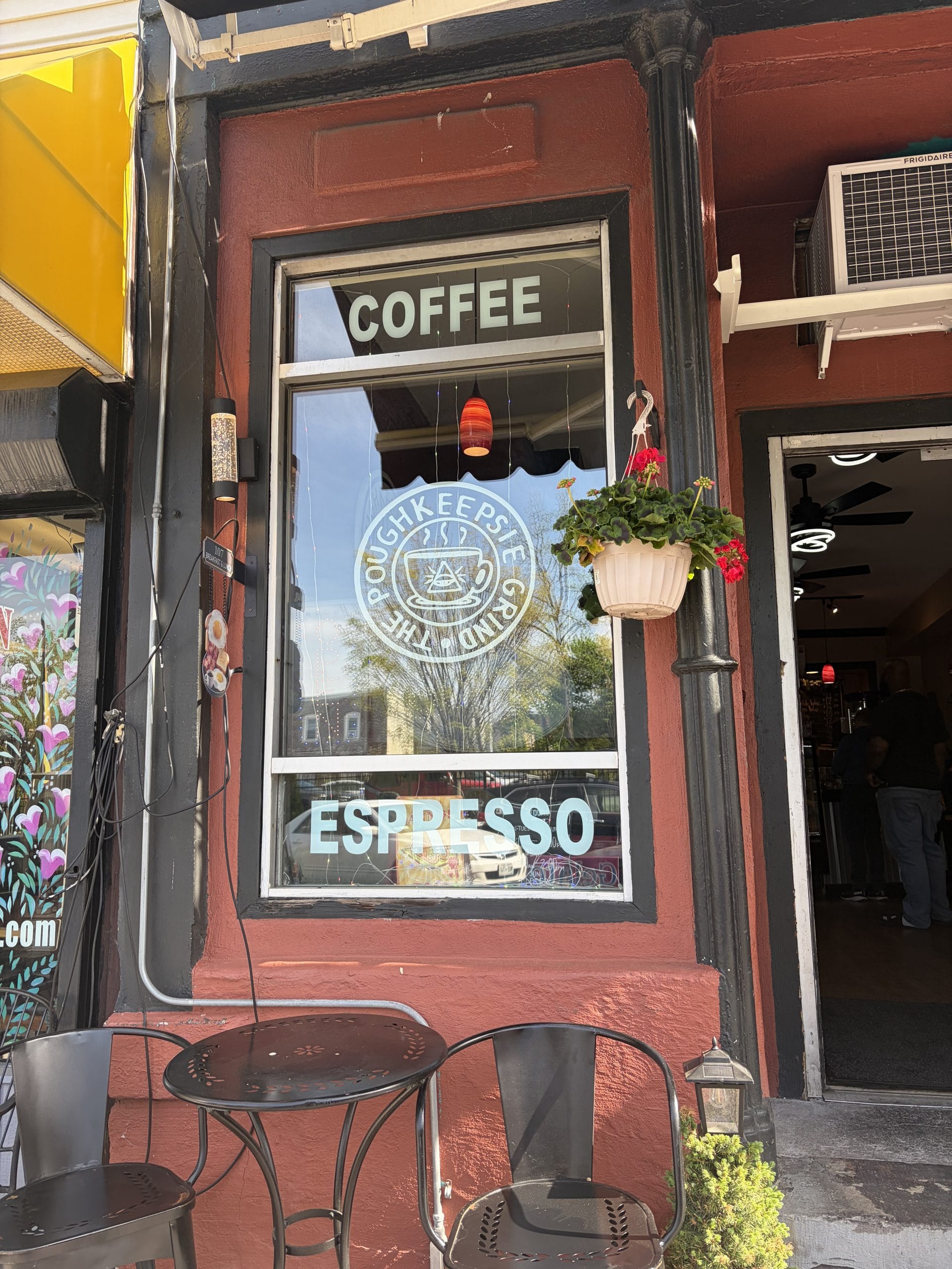 A coffee shop window with Coffee and Espresso written on the glass, featuring a round logo—perfect for a pause during your Walking Tour of Poughkeepsie. Two metal chairs and a small table sit outside, with a hanging flower pot beside the door.