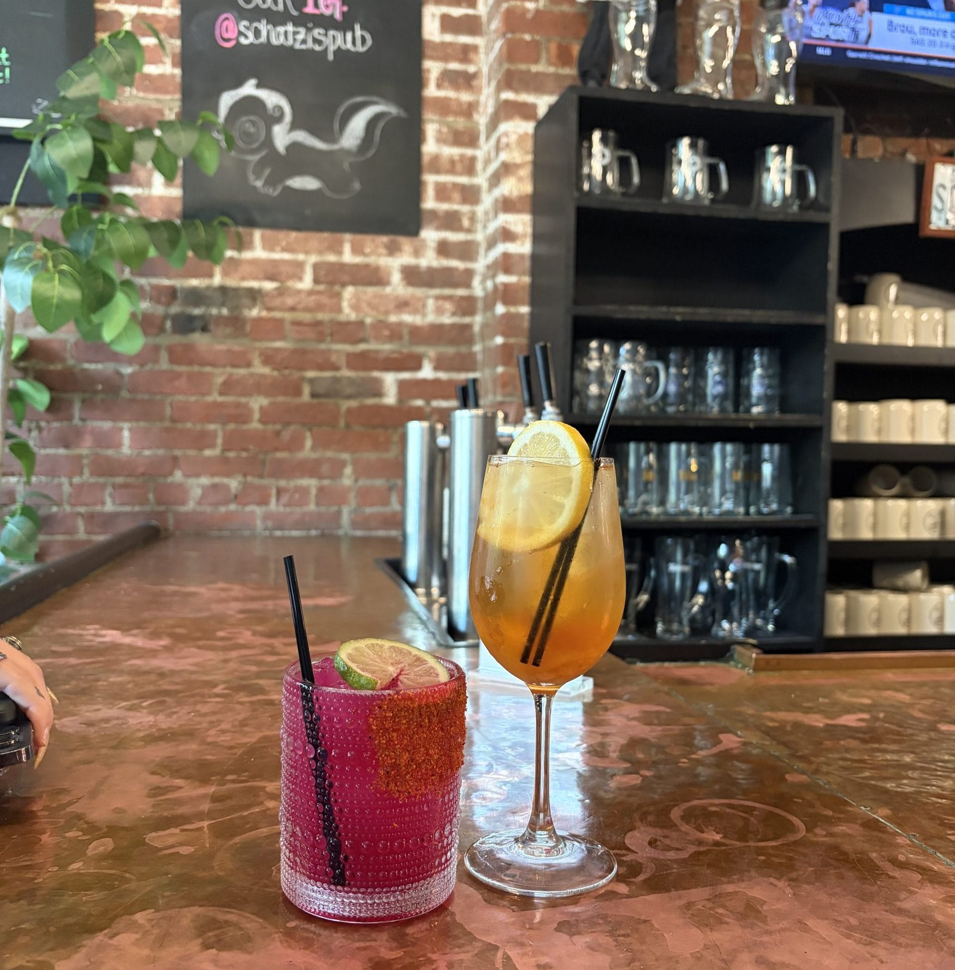 A copper bar top with two cocktails—one in a short glass with lime and strawberry, the other in a wine glass with an orange drink and lemon slice—awaits after a Walking Tour of Poughkeepsie. A chalkboard menu, mugs, and TV complete the cozy scene.