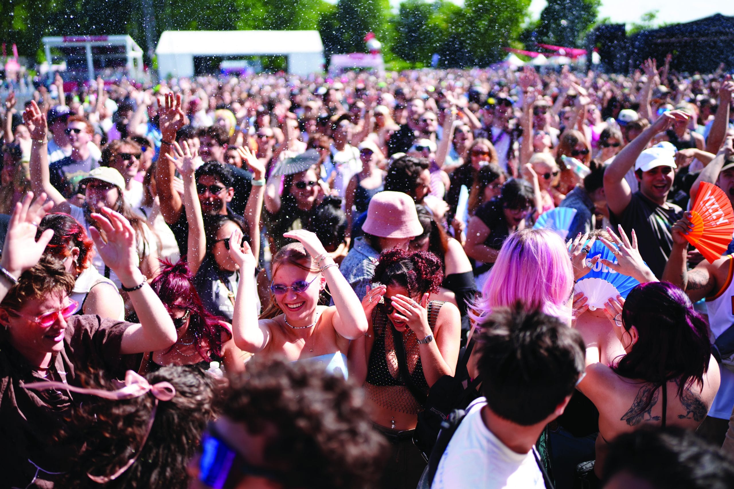 A large crowd of people out of doors at a festival, many smiling and dancing with arms raised. The sun is shining, and some enjoy true great adventures with fans or sunglasses. Trees and tents fill the vibrant background.