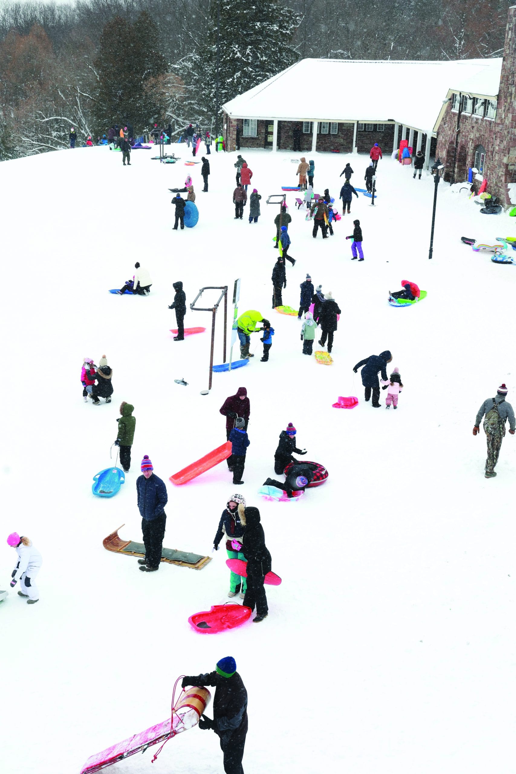 Many people, including children and adults, are enjoying outdoor adventures as they sled and play in the snow near a building. Colorful sleds are scattered across the snowy ground on a lively winter day.