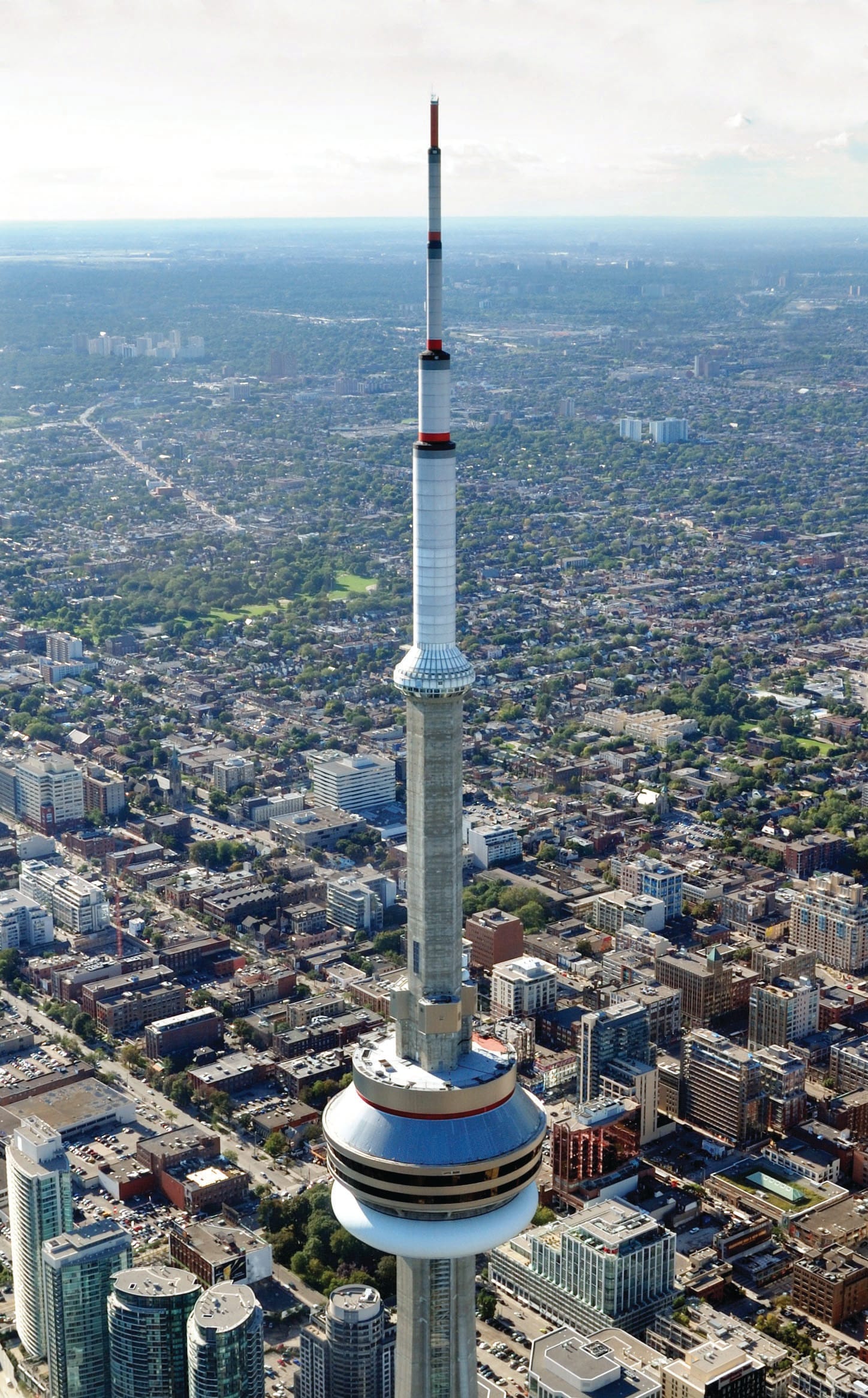 Aerial view of the CN Tower rising above downtown Toronto, where city buildings, roads, and greenery set the scene for power walking enthusiasts under a partly cloudy sky.