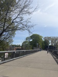 A wide pedestrian bridge with metal railings, bordered by trees with spring foliage under a partly cloudy sky. A single person walks ahead near the middle distance, evoking a peaceful Walking Tour of Poughkeepsie, with buildings visible to the left.