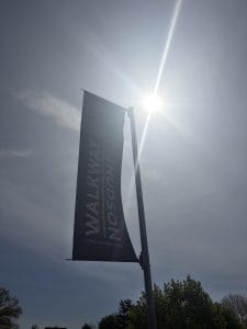 A tall banner reading WALKWAY OVER THE HUDSON STATE HISTORIC PARK hangs from a pole, silhouetted against a bright sun and a partly cloudy sky, with green treetops below—an inviting stop on any Walking Tour of Poughkeepsie.