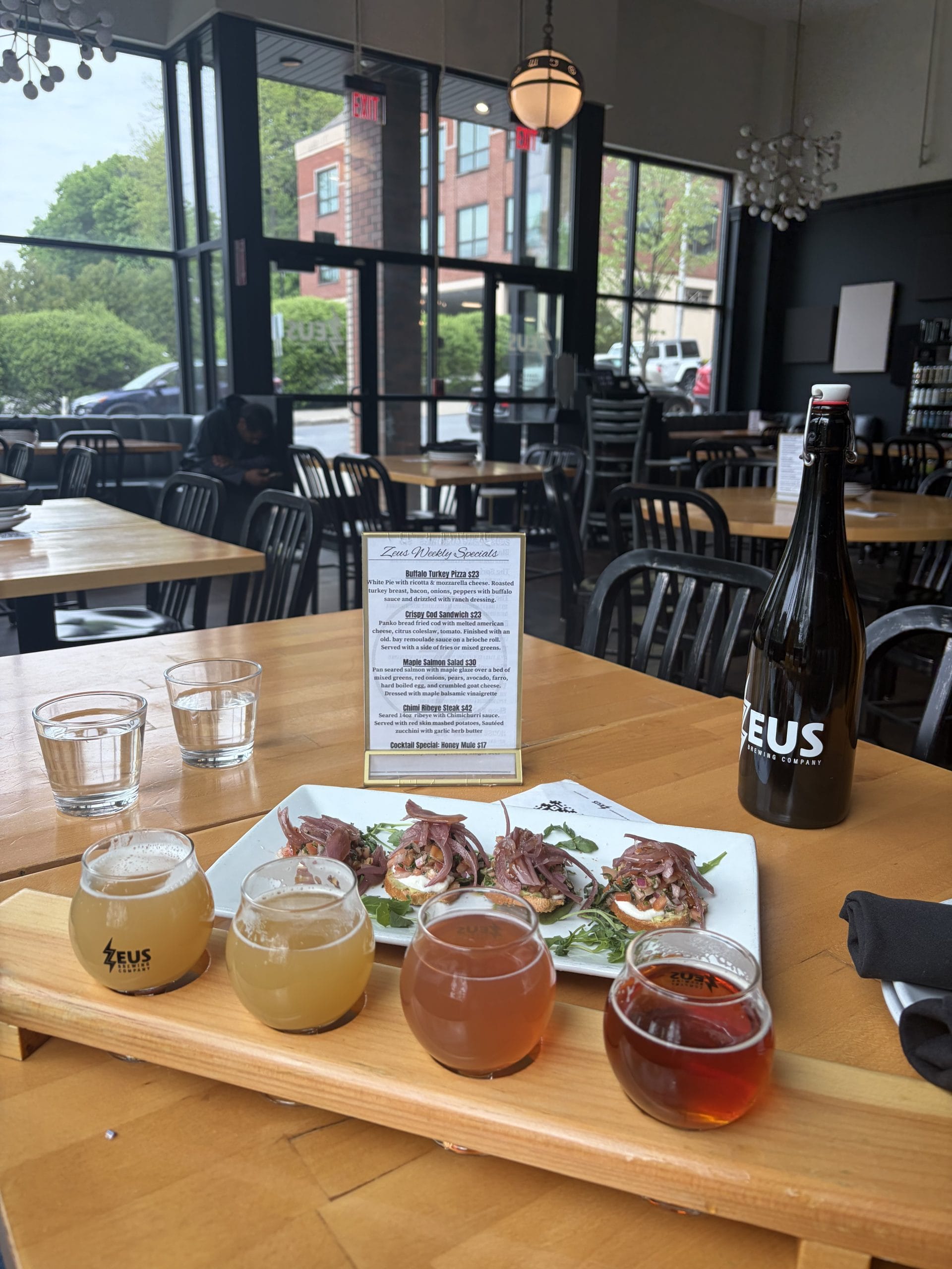 A table at a restaurant during a Walking Tour of Poughkeepsie showcases four small glasses of beer, a ZEUS-labeled bottle, appetizers topped with meat, two waters, and a menu. Large windows frame lush greenery and a brick building outside.