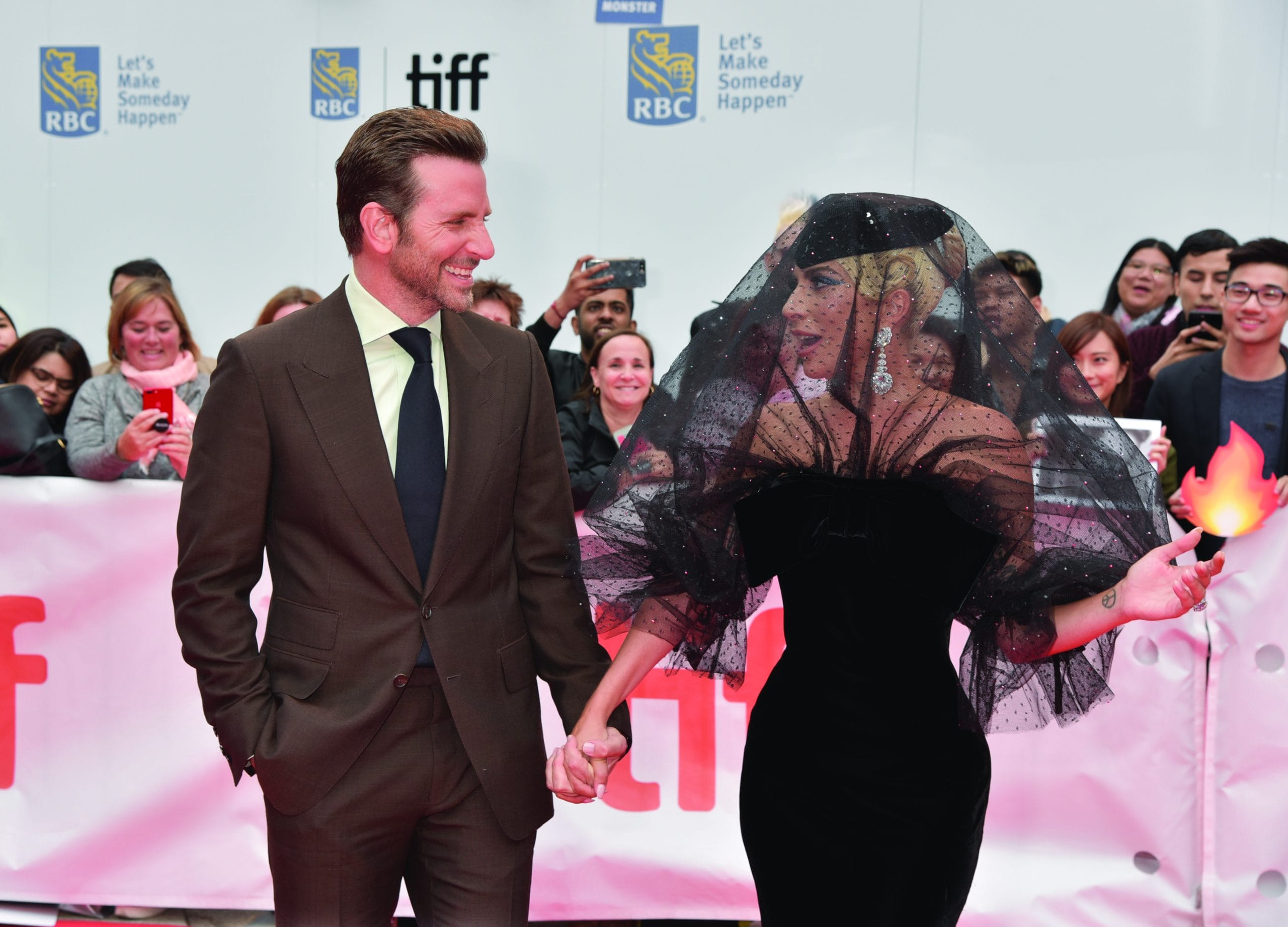 A man in a brown suit and a woman in a dramatic black dress with a sheer veil hold hands and smile at each other on the red carpet, capturing the glamour and excitement of the Toronto arts scene amid fans and photographers.